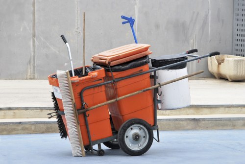 A mobile rubbish collection trolley with large black rubber wheels, positioned outdoors on a paved surface with a concrete wall in the background. The trolley is primarily orange with a black storage compartment and holds various cleaning and waste collection tools, including a large sweeping brush with black bristles, a long-handled tool with a blue grip, and a beige foam float. A folded orange mat rests on top of the trolley, while a white container and a black hose are attached on the side. The scene suggests a commercial waste removal or cleaning service setup, possibly for a premises or outdoor area within Chelmsford, with the trolley ready for debris collection or street cleaning tasks conducted by a company like Commercial Waste Chelmsford.