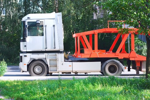 A side view of a commercial flatbed truck parked on a paved surface, featuring a white cab with a large windshield and side mirror, and an orange metal frame structure at the back designed for transporting waste or bulky rubbish. The flatbed area is currently empty, with visible mechanical components and attachment points. The vehicle is positioned in an outdoor setting with a background of leafy trees and greenery, suggesting a residential or commercial area. The scene is well-lit with natural daylight, highlighting the clean and maintained appearance of the truck, used in waste collection services such as those offered by Commercial Waste Chelmsford in the town. The image emphasizes the equipment used for rubbish removal and waste hauling, relevant to services in Chelmsford's postcode region.