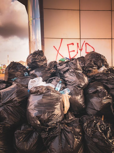 A metal waste disposal station positioned on a train station platform, featuring four separate color-coded bins for different types of rubbish. The first bin on the left, marked with a yellow label, i
